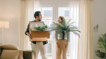 Two people stand in an apartment holding housplants